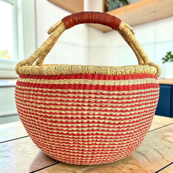 XL round Bolga basket with fine red and natural stripes and a leather-wrapped handle, on a wooden table