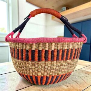 XL round Bolga basket with upright orange and black stripes, a pink rim and a leather-wrapped handle, on a wooden table