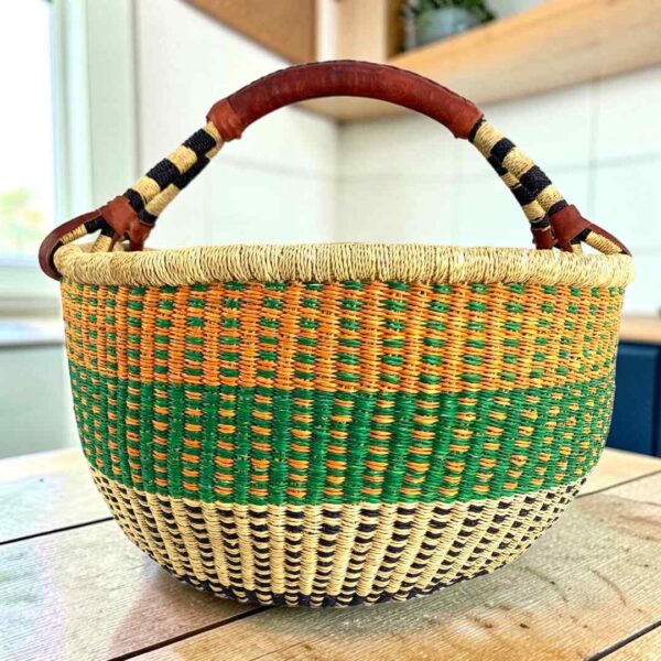 XL round Bolga basket with orange and green checks over a natural base and a striped leather handle, on a wooden table
