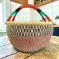 XL round Bolga basket with black checked bands over a red and green speckled weave and a striped leather handle, on a wooden table