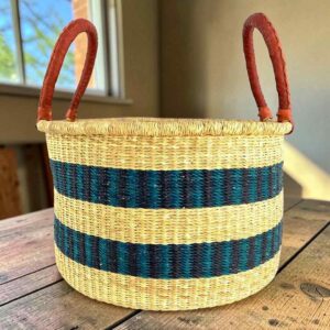 Hamper basket in natural straw with two dark-toned stripes and leather handles, on a wood table