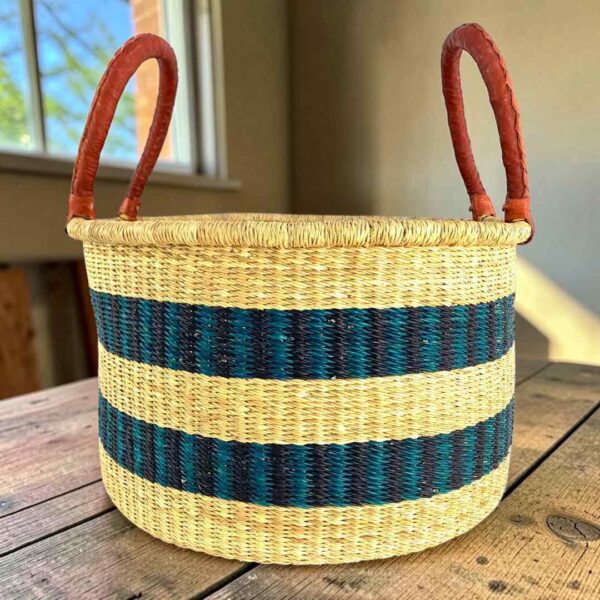 hamper-basket-set-028-pickleball-natural-dark-stripes-woven-bashiri-main Hamper basket in natural straw with two dark-toned stripes and leather handles, on a wood table