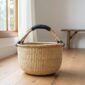 a large bolga basket, woven from elephant grass, standing on a wooden surface, with a black handle