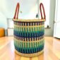 Open laundry basket with navy, teal and green bands on natural straw and tall leather handles