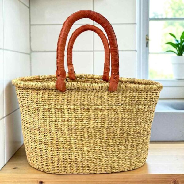 Oval Bolga basket with natural undyed straw and leather handles, on a wooden kitchen bench