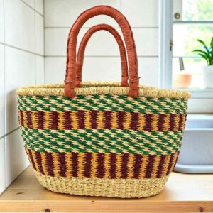 Oval Bolga basket with a green checkered band over maroon and gold stripes and leather handles, on a wooden kitchen bench