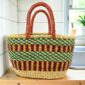 Oval Bolga basket with a green checkered band over maroon and gold stripes and leather handles, on a wooden kitchen bench