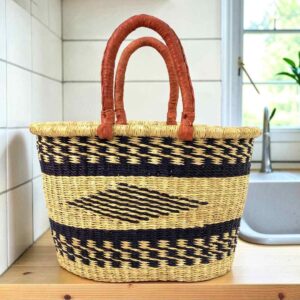 Oval Bolga basket with black bands and a central black diamond motif and leather handles, on a wooden kitchen bench