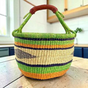 Large round Bolga basket with bright leafy and citrus bands and deep inky stripes, leather-wrapped handle, on a timber benchtop
