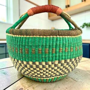 Large round Bolga basket with a fresh leafy band over a warm earthy checkerboard, leather-wrapped handle, on a timber benchtop