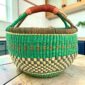 Large round Bolga basket with a fresh leafy band over a warm earthy checkerboard, leather-wrapped handle, on a timber benchtop