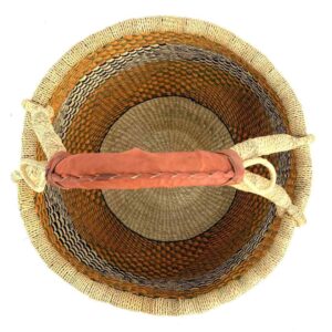 Top-down view inside the large round Bolga basket with warm amber tones and a dark diagonal checkerboard, showing the coiled base and leather handle