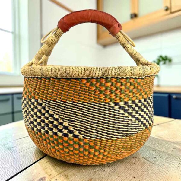Large round Bolga basket with warm amber tones and a dark diagonal checkerboard, leather-wrapped handle, on a timber benchtop