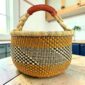 Large round Bolga basket with warm amber tones and a dark diagonal checkerboard, leather-wrapped handle, on a timber benchtop