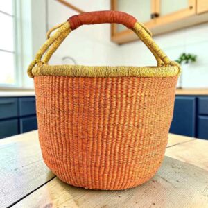 Large round Bolga basket with a single warm terracotta tone and natural rim, leather-wrapped handle, on a timber benchtop