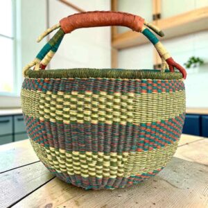 Large round Bolga basket with muted olive and slate tones with natural checks, leather-wrapped handle, on a timber benchtop