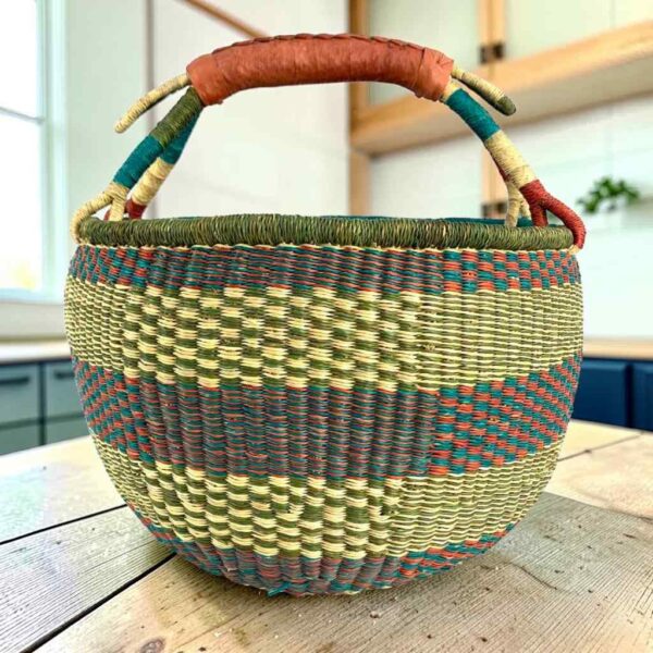 Large round Bolga basket with muted olive and slate tones with natural checks, leather-wrapped handle, on a timber benchtop