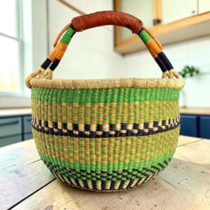 Large round Bolga basket with fresh leafy tones and a band of deep inky checks, leather-wrapped handle, on a timber benchtop