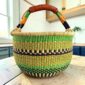 Large round Bolga basket with fresh leafy tones and a band of deep inky checks, leather-wrapped handle, on a timber benchtop