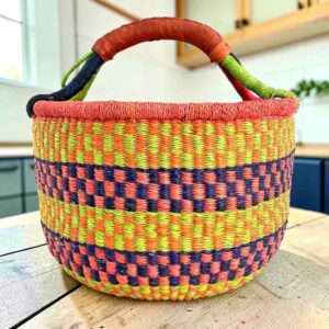 Large round Bolga basket with bright citrus and rosy bands with deep inky checks, leather-wrapped handle, on a timber benchtop