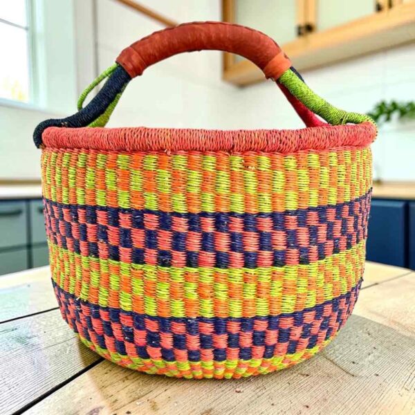 Large round Bolga basket with bright citrus and rosy bands with deep inky checks, leather-wrapped handle, on a timber benchtop