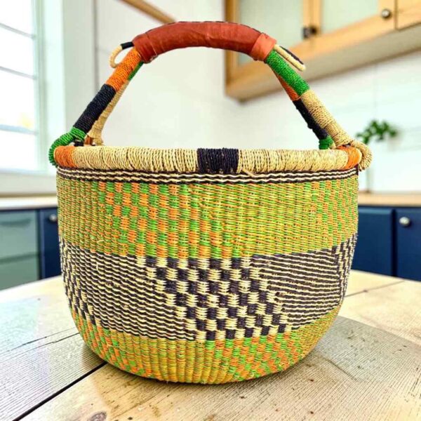 Large round Bolga basket with zesty citrus tones and a diagonal inky checked section, leather-wrapped handle, on a timber benchtop