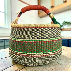 Large round Bolga basket with fine dark checks and rust and leafy stripes, leather-wrapped handle, on a timber benchtop