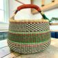 Large round Bolga basket with fine dark checks and rust and leafy stripes, leather-wrapped handle, on a timber benchtop