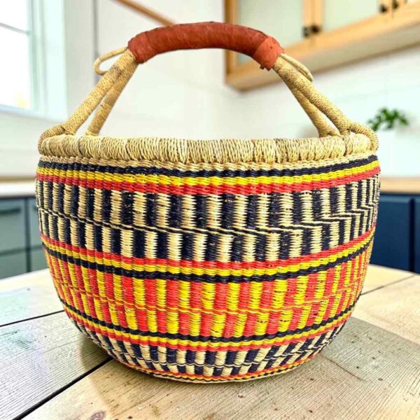 Large round Bolga basket with sunny and fiery bands with dark zigzag rows, leather-wrapped handle, on a timber benchtop