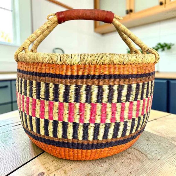 Large round Bolga basket with warm burnt tones framing rosy and inky stripes, leather-wrapped handle, on a timber benchtop