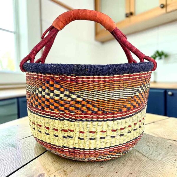 Large round Bolga basket with an inky rim over rust and amber patterning, leather-wrapped handle, on a timber benchtop