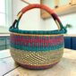 Large round Bolga basket with deep sea-green bands and berry stripes, leather-wrapped handle, on a timber benchtop