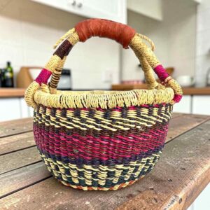 Small round Bolga basket with pink and navy bands on natural straw and a leather-wrapped handle, on a wooden table