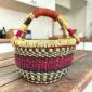 Small round Bolga basket with pink and navy bands on natural straw and a leather-wrapped handle, on a wooden table