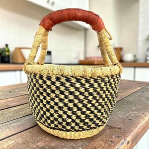 Small round Bolga basket with a black and natural checked weave and a leather-wrapped handle, on a wooden table