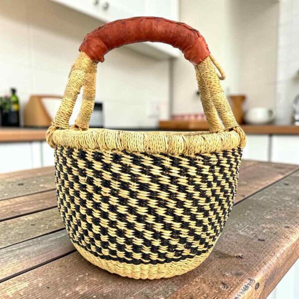 Small round Bolga basket with a black and natural checked weave and a leather-wrapped handle, on a wooden table
