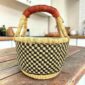 Small round Bolga basket with a black and natural checked weave and a leather-wrapped handle, on a wooden table
