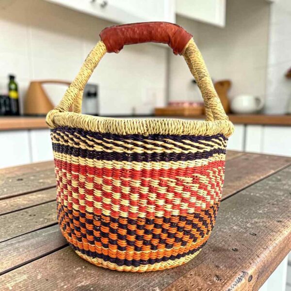Small round Bolga basket with orange, red and navy stacked bands and a leather-wrapped handle, on a wooden table