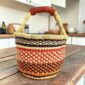 Small round Bolga basket with orange, red and navy stacked bands and a leather-wrapped handle, on a wooden table