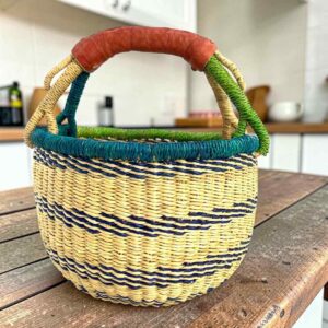 Small round Bolga basket with navy stripes on natural straw with a teal and green rim and a leather-wrapped handle, on a wooden table