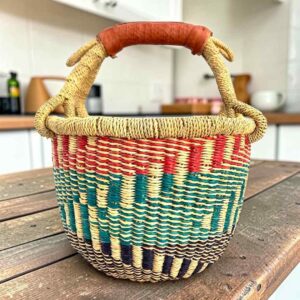 Small round Bolga basket with teal, red and dark stepped bands and a leather-wrapped handle, on a wooden table