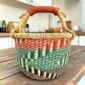 Small round Bolga basket with teal, red and dark stepped bands and a leather-wrapped handle, on a wooden table