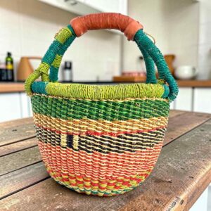 Small round Bolga basket with green rim and coral, orange and dark bands and a leather-wrapped handle, on a wooden table