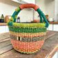 Small round Bolga basket with green rim and coral, orange and dark bands and a leather-wrapped handle, on a wooden table