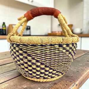 Small round Bolga basket with a navy and natural staggered check and a leather-wrapped handle, on a wooden table
