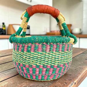 Small round Bolga basket with teal and pink checked bands with a teal rim and a leather-wrapped handle, on a wooden table