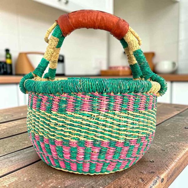 Small round Bolga basket with teal and pink checked bands with a teal rim and a leather-wrapped handle, on a wooden table