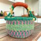 Small round Bolga basket with teal and pink checked bands with a teal rim and a leather-wrapped handle, on a wooden table