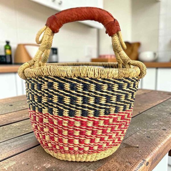 Small round Bolga basket with navy checks over red and pink bands and a leather-wrapped handle, on a wooden table