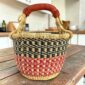 Small round Bolga basket with navy checks over red and pink bands and a leather-wrapped handle, on a wooden table
